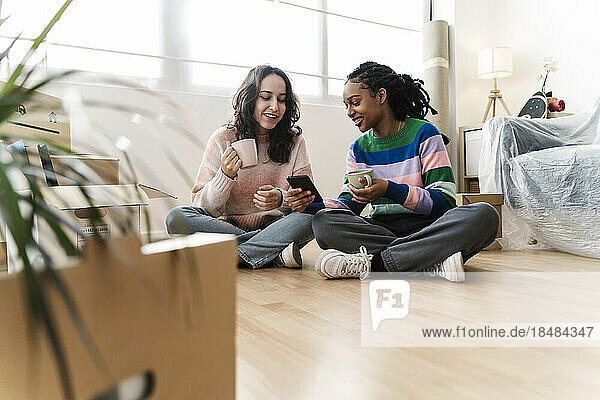 Lächelnde junge Frau  die zu Hause auf dem Boden sitzt und ihr Smartphone teilt