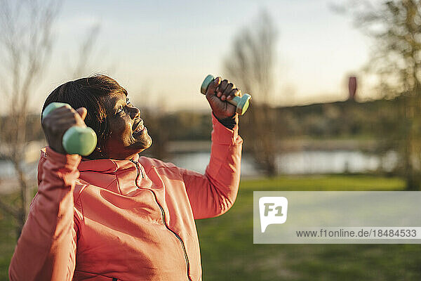 Senior woman holding dumbbells exercising in park at sunset