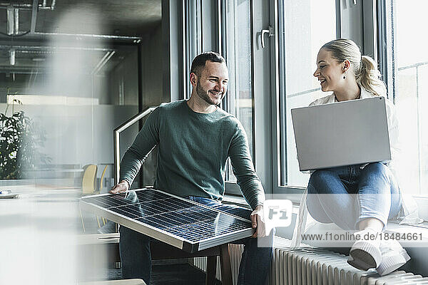 Smiling businessman holding solar panel with colleague sitting at office