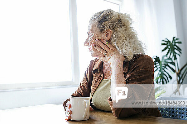 Lonely senior woman with mug sitting by table at home
