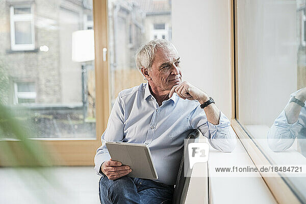 Thoughtful senior businessman with tablet PC sitting near window at office