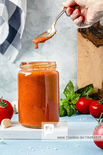 Hand of woman holding spoon with tomato sauce over jar on table