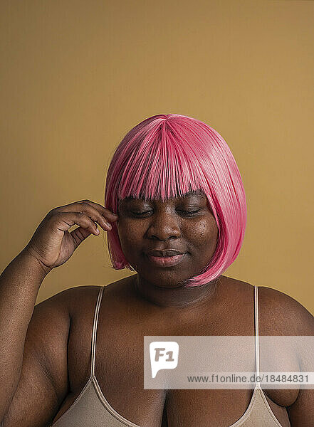 Plus size woman wearing pink bob wig posing with eyes closed against beige background