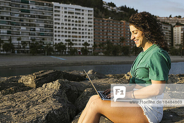 Smiling freelancer using laptop sitting on rock at beach