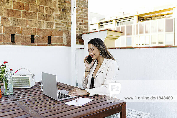 Smiling freelancer talking on smart phone and using laptop at rooftop