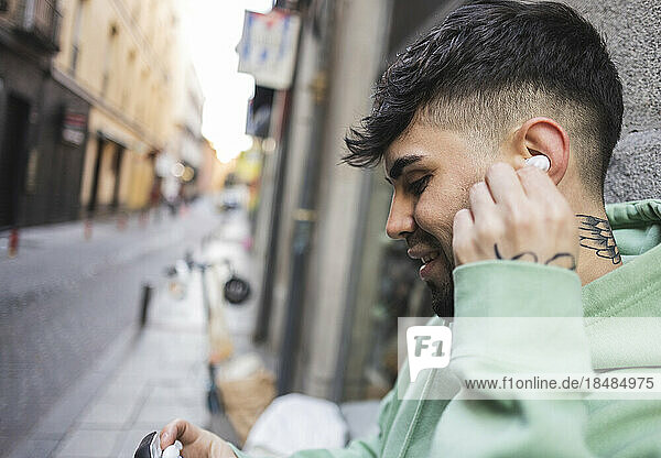 Smiling man putting in earphone by wall