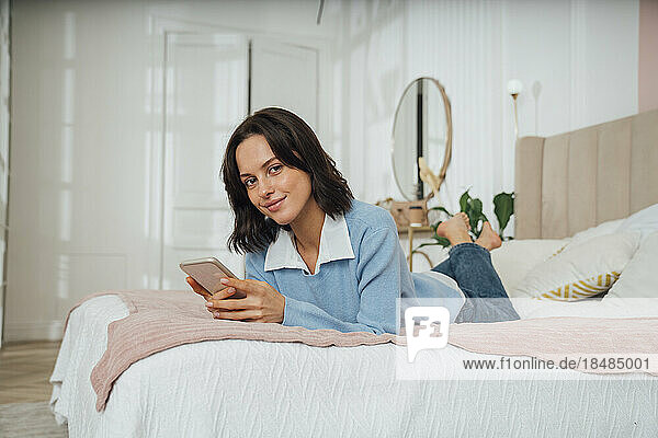 Smiling young woman lying on bed with smart phone at home
