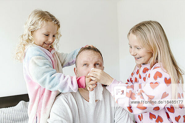 Happy daughters wiping nose of father with napkin at home
