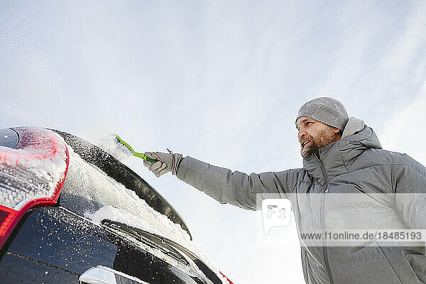 Man wearing knit hat brushing snow from car
