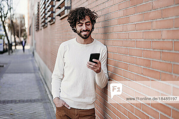 Happy young man using smart phone leaning on wall