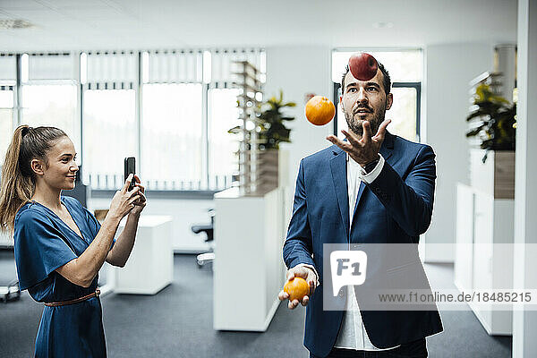 Businesswoman taking photo using smart phone of colleague juggling fruits in office