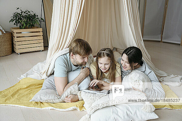 Smiling parents and daughter reading book in blanket tent