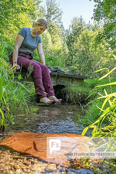 Frau sitzt an Bach und hält die Füße in Wasser auf dem Wanderweg Sprollenhäuser Hut  Bad Wildbad  Schwarzwald  Deutschland  Europa