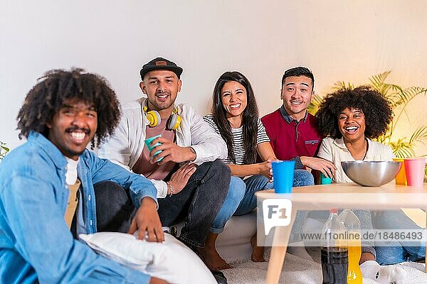 Gruppenporträt multiethnischer Freunde  die auf dem Sofa Popcorn essen und Limonade trinken und lächelnd in die Kamera schauen