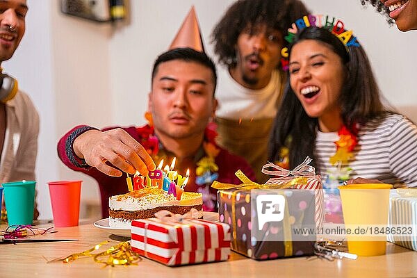 Multiethnische Gruppe von Freunden bei einer Geburtstagsfeier auf dem Sofa zu Hause mit einem Kuchen und Geschenken  chinesischer Mann zündet die Kerzen an