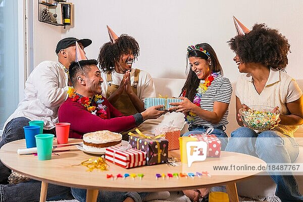 Gruppe von Freunden bei einer Geburtstagsfeier auf dem Sofa zu Hause  die lächelnd die Geschenke überreichen und das Geburtstagskind überraschen