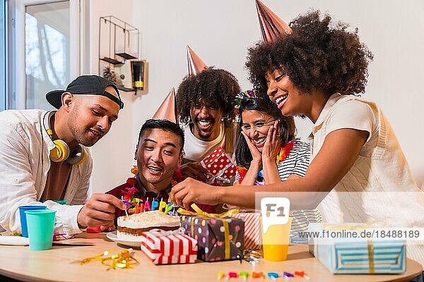 Multiethnische Gruppe von Freunden bei einer Geburtstagsfeier auf dem Sofa zu Hause mit einem Kuchen und Geschenken  lächelnd Platzierung der Kerzen