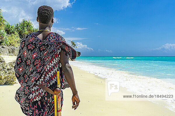 Maasai-Mann mit traditioneller Kleidung und Stock bewundert das kristallklare Meer am Strand  Sansibar  Tansania  Ostafrika  Afrika