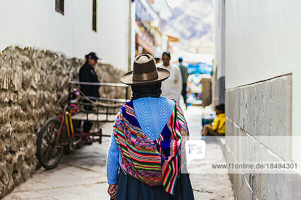Woman walking away in narrow street with traditional colorful Peruvian bag over her back  Pisaq  Peru  South America