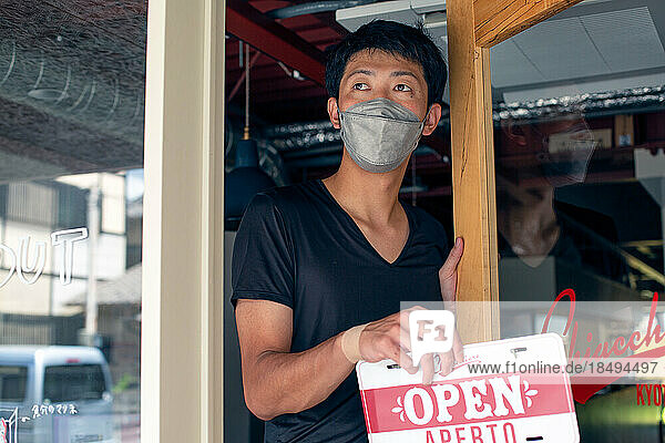 Man wearing a face mask hanging up an Open sign on an Italian restaurant door. Dual language  Italian and English.