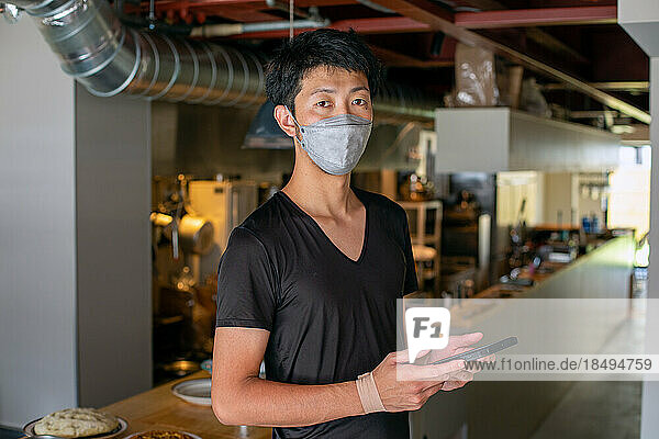 A man working in a restaurant  wearing a face mask  by an open kitchen  holding a digital tablet.