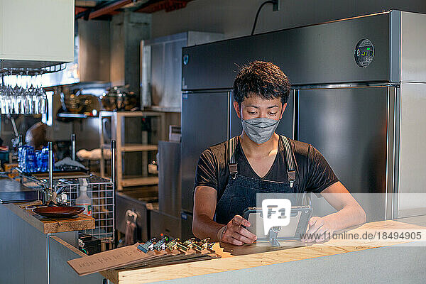 A man wearing a face mask at the counter of a restaurant kitchen  using a digital tablet  the owner or manager.