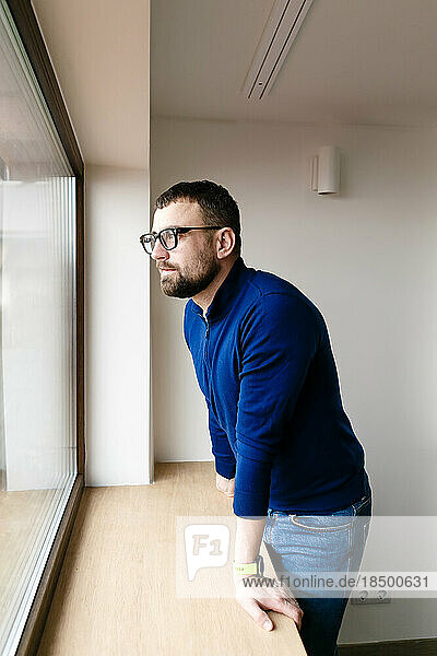 brunette man in glasses and a blue sweater looks out the window alone