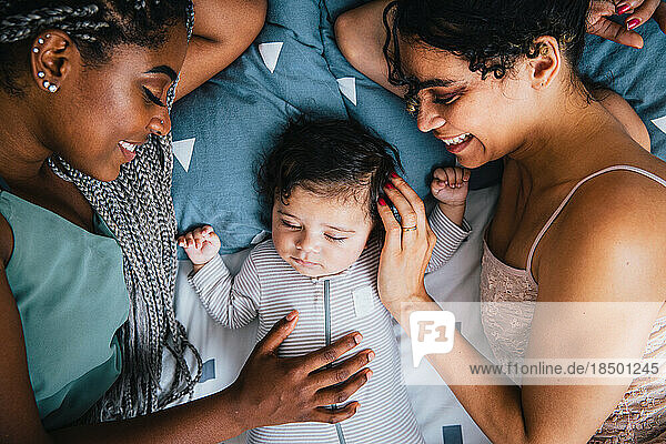 Overhead view of happy girlfriends with cute sleeping baby boy resting on bed