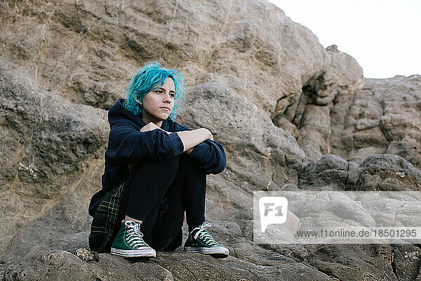 Pensive teen girl with blue hair sits on large rock
