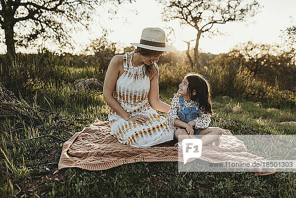 Mother and young daughter sitting on blanket in backlit meadow