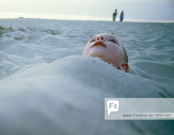 A young boy buried in sand on the beach.