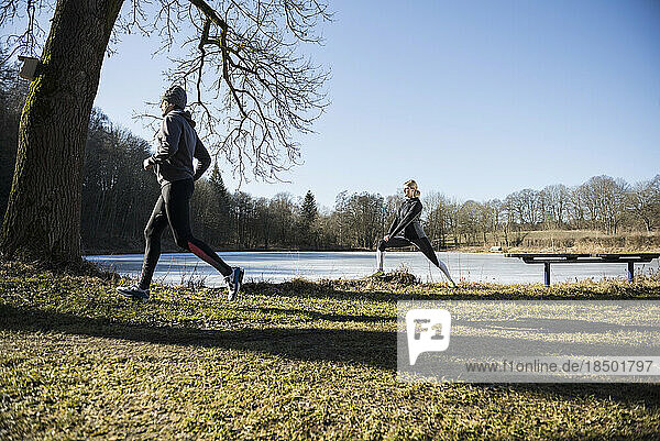 Man jogging while woman stretching on fitness trail in front of frozen lake