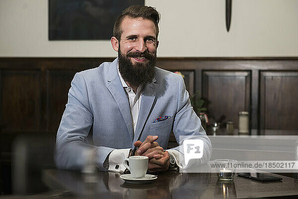 Portrait of smart man sitting by table with coffee cup and mobile phone on it at restaurant
