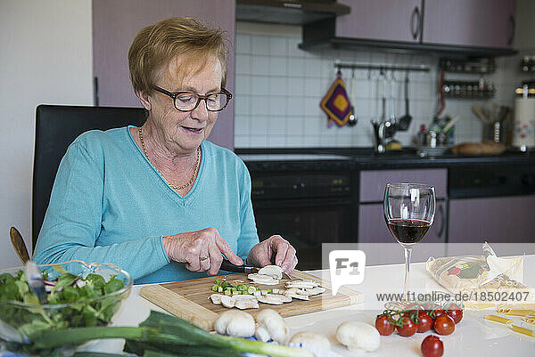 Old woman cutting spring onions and button mushroom at the kitchen table