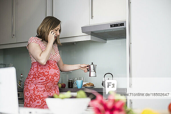 Pregnant woman talking on smartphone and making coffee for breakfast in the kitchen  Munich  Bavaria  Germany