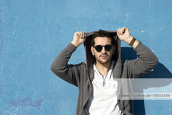 Young bearded man leaning on blue wall wearing sunglasses