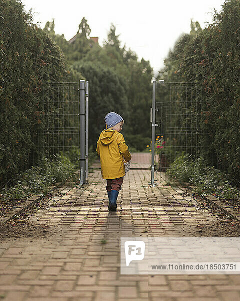 Small boy in yellow raincoat walking away on pavement in garden