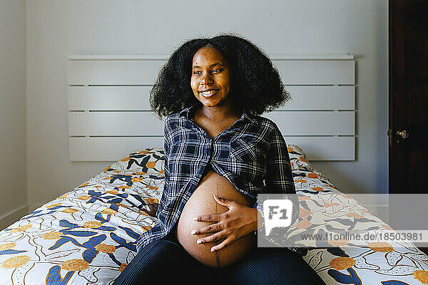 A young woman sits on bed holding pregnant belly