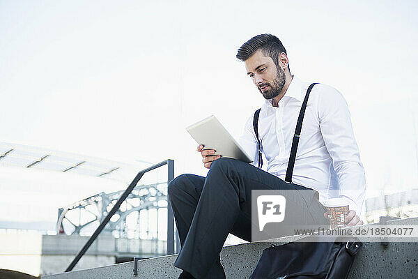 Businessman sitting on the stairs and working on digital tablet with coffee  Munich  Bavaria  Germany