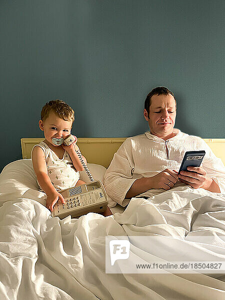 Father and son with pacifier hold telephone on bed