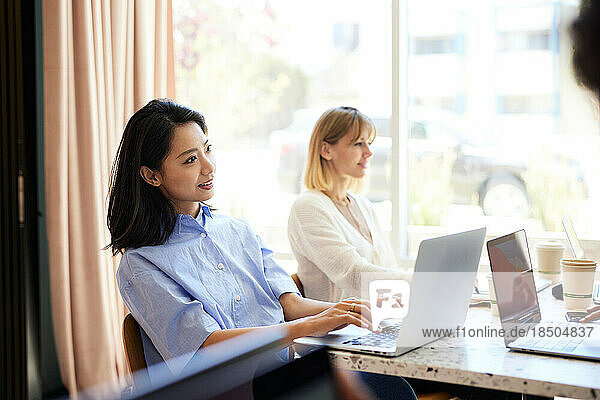 Smiling businesswoman using laptop sitting by colleague at cafe