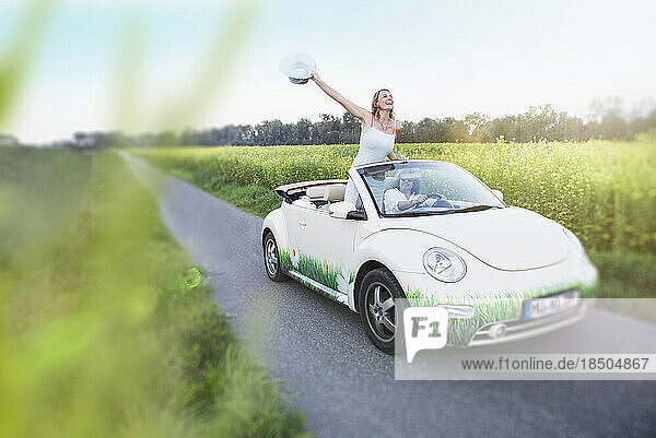 Happy couple driving in a car Beetle Cabrio woman waving with hat  Bavaria  Germany