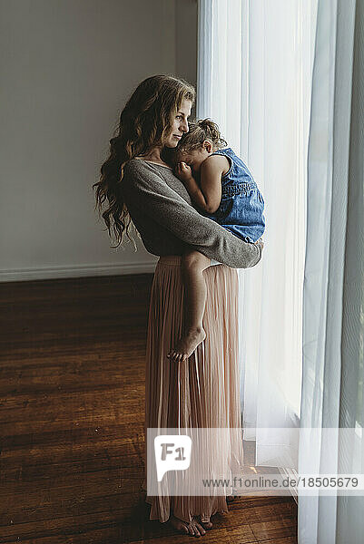 Full length view of mother and daughter cuddling by window in studio