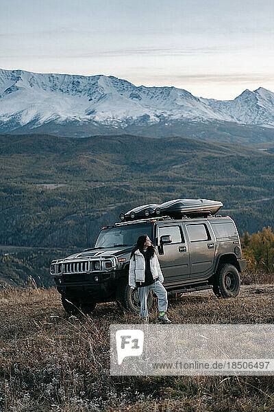 A girl stands next to a black SUV against the background of a mountain