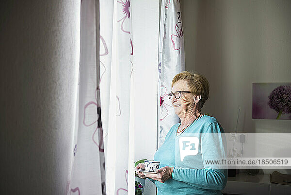 Smiling old woman looking out of window with a cup of coffee