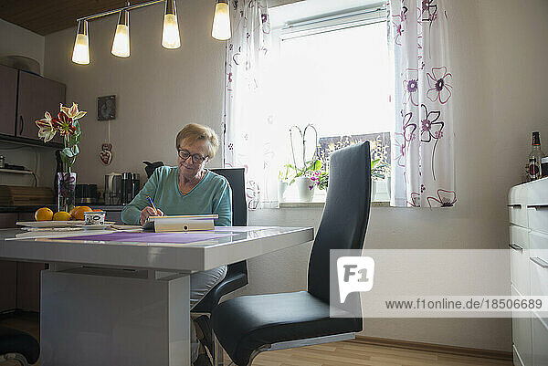 Old woman watching digital tablet and writing in book at dining table