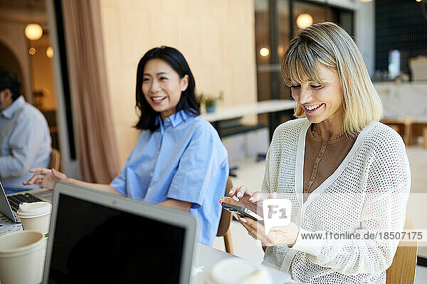 Happy businesswoman using mobile phone by female colleague at cafe