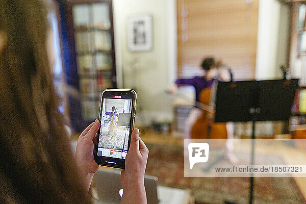 A child with cellphone records brother playing cello at home