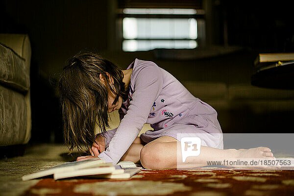 A small child sits on rug in morning light with pile of books reading