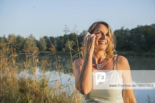 Cheerful woman smiling while talking on smart phone at riverbank  Bavaria  Germany